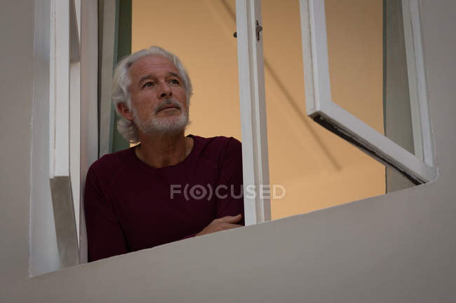 Homem idoso atencioso olhando através da janela em casa — Fotografia de Stock