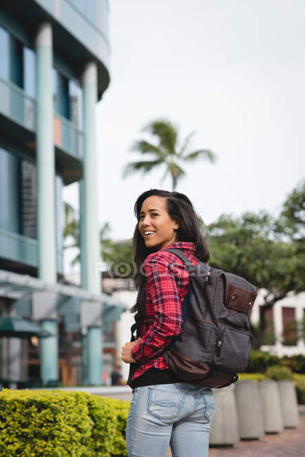 Smiling woman with backpack standing on street and looking over shoulder — Stock Photo