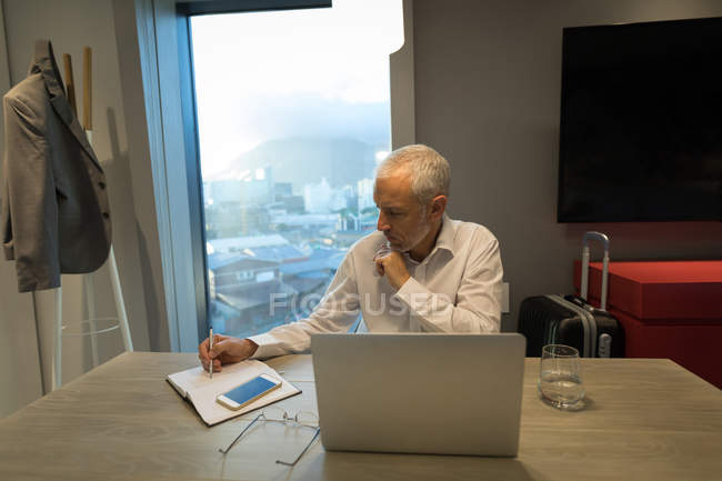 Empresario escribiendo notas en el diario en el escritorio en la habitación del hotel - foto de stock