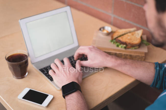 Gros plan de l'homme utilisant un ordinateur portable dans un café avec un sandwich sur la table . — Photo de stock