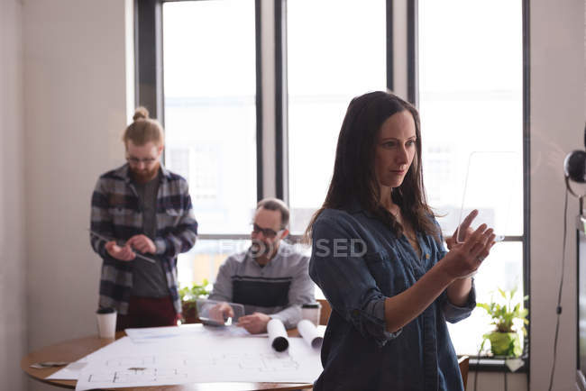 Жіночий керівник використовує скляний цифровий планшет в офісі з колегами, що працюють у фоновому режимі — стокове фото