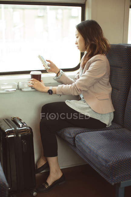 Жінка-виконавця, використовуючи мобільний, маючи каву в поїзді — стокове фото