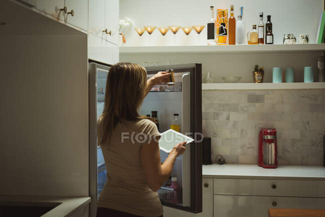 Femme utilisant une tablette numérique tout en ouvrant un réfrigérateur à la maison — Photo de stock