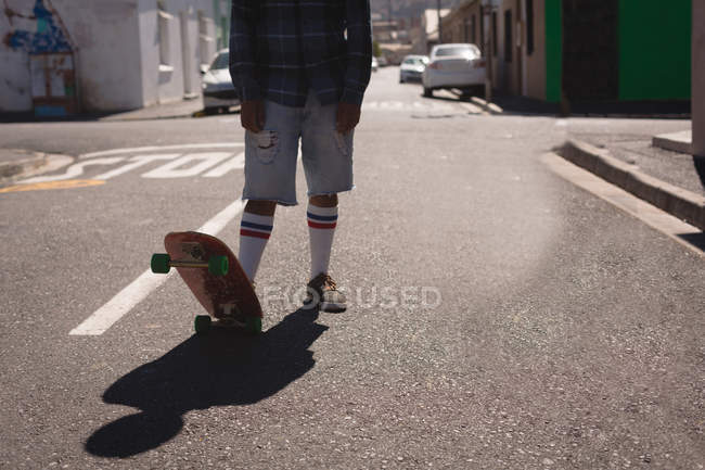 Section basse de l'homme debout avec planche à roulettes sur la rue en plein soleil — Photo de stock