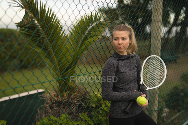 Mujer reflexiva sosteniendo raqueta y pelota de tenis en pista de tenis - foto de stock