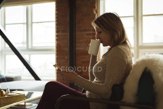 Donna che usa il telefono cellulare mentre prende il caffè a casa — Foto stock