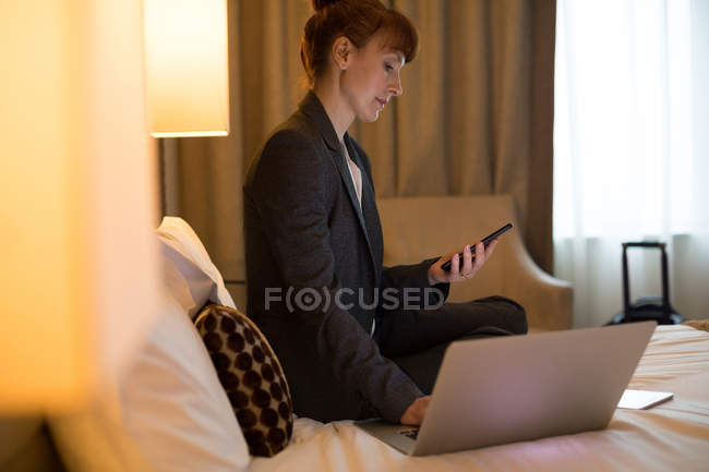 Деловая женщина с помощью мобильного телефона во время работы на ноутбуке в номере отеля — стоковое фото