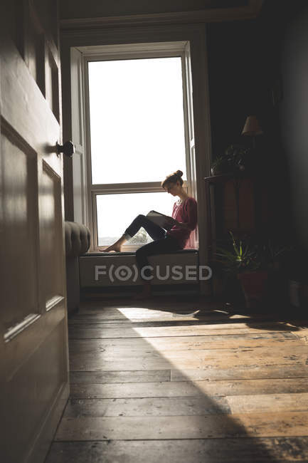 Frau liest Buch auf Fensterbank zu Hause — Stockfoto