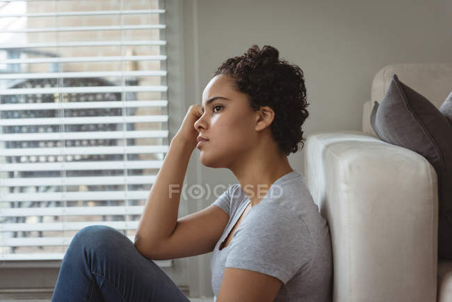 Mujer pensativa sentada cerca del sofá en casa - foto de stock