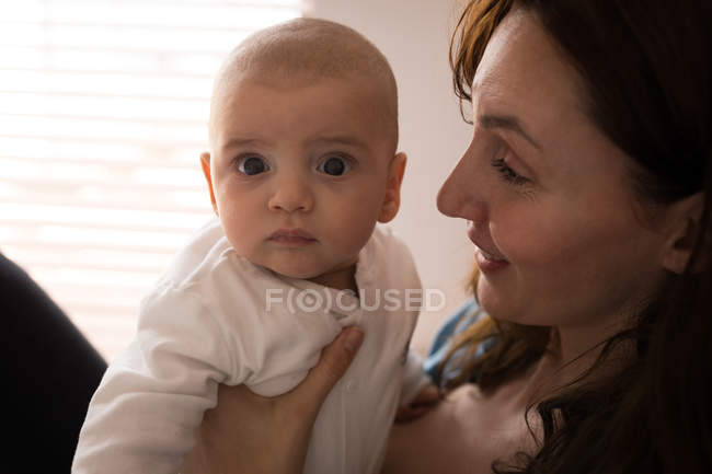 Retrato de lindo bebé mirando a la cámara y mamá sosteniéndolo en casa - foto de stock