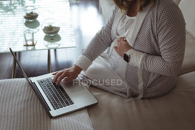 Schwangere benutzt Laptop, während sie ihren Bauch zu Hause berührt — Stockfoto