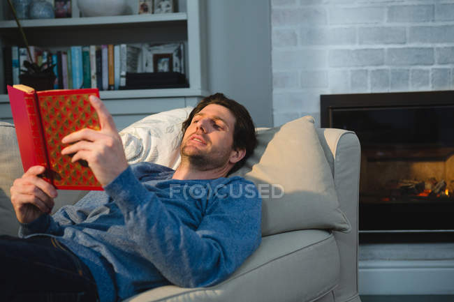 Homme lisant un livre alors qu'il était allongé sur le canapé dans le salon à la maison — Photo de stock