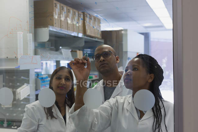 Científicos escribiendo nota en pared de vidrio en el laboratorio - foto de stock