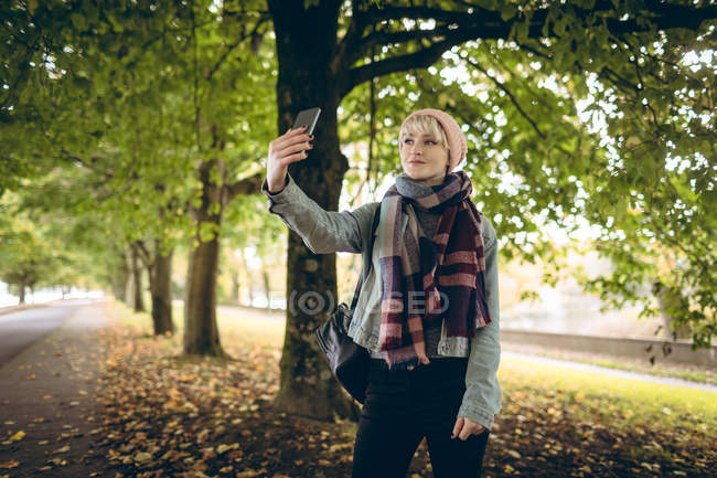 Молодая женщина в теплой одежде щёлкает селфи в парке — стоковое фото