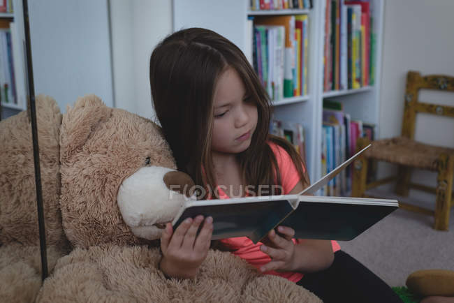 Niño de edad elemental con libro de lectura de oso de peluche en la sala de estar en casa - foto de stock