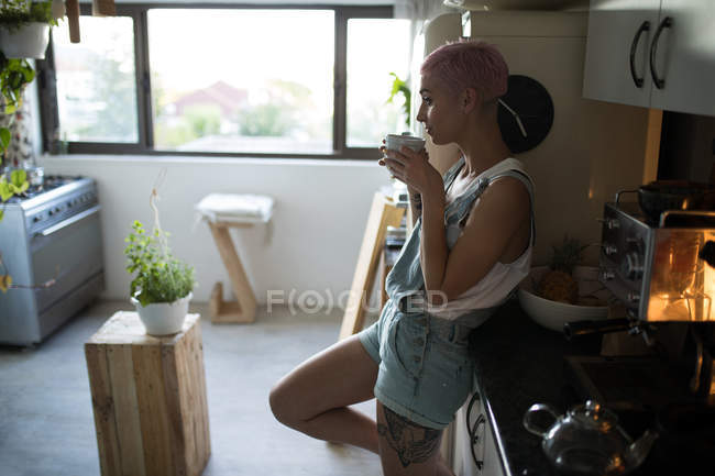 Mulher elegante com cabelo rosa bebendo café na cozinha em casa . — Fotografia de Stock