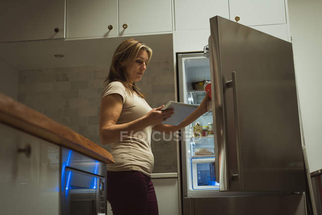 Жінка використовує цифровий планшет під час відкриття холодильника вдома — стокове фото