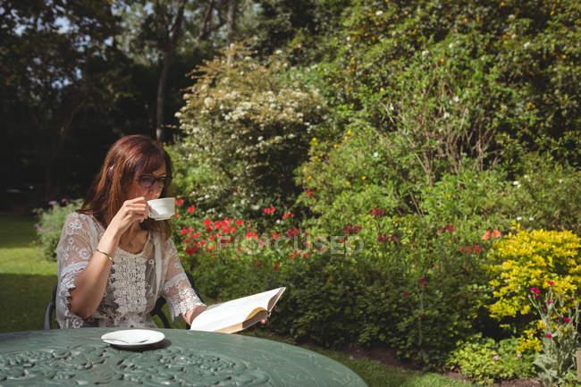 Жінка читає книгу, маючи каву в саду — стокове фото