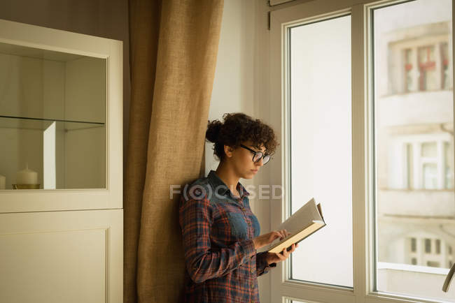 Mujer leyendo libro cerca de ventana en casa - foto de stock