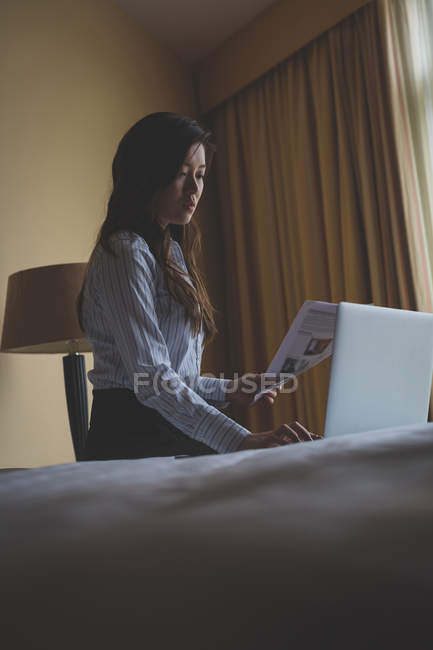 Empresária segurando documentos enquanto trabalhava em laptop no quarto de hotel — Fotografia de Stock
