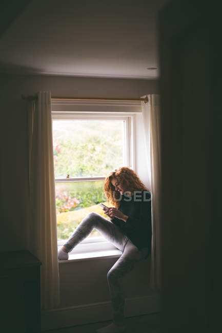 Mulher usando telefone celular enquanto sentado no peitoril da janela em casa — Fotografia de Stock