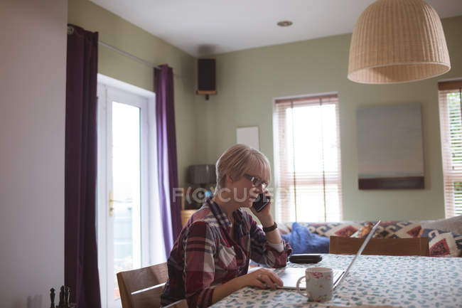 Зріла жінка розмовляє на мобільному телефоні, використовуючи ноутбук вдома — стокове фото