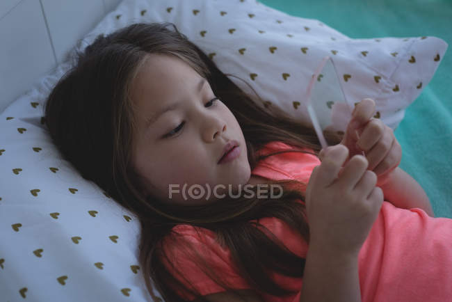 Niña de edad elemental utilizando el teléfono móvil de vidrio en la cama en casa - foto de stock