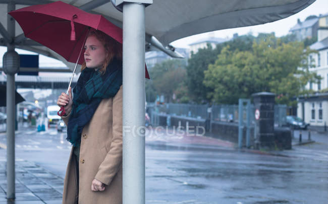Junge Frau mit Regenschirm steht an einem regnerischen Tag an einer Bushaltestelle — Stockfoto