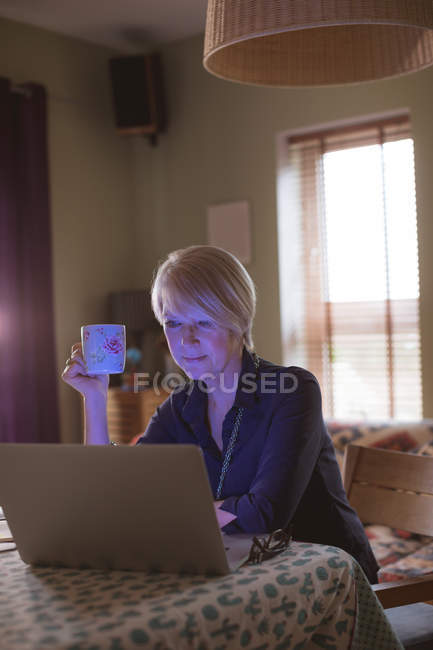 Mulher madura usando laptop enquanto toma café na sala de estar em casa — Fotografia de Stock