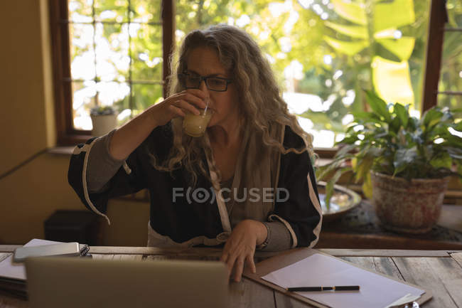 Femme mature travaillant sur un ordinateur portable tout en buvant du jus à la maison — Photo de stock
