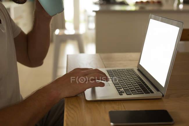 Средняя секция человека, пьющего кофе во время использования ноутбука дома — стоковое фото