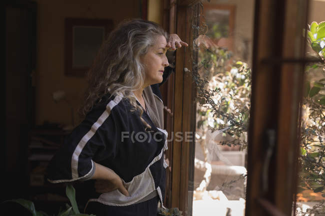 Задумчивая зрелая женщина смотрит в окно на дом — стоковое фото