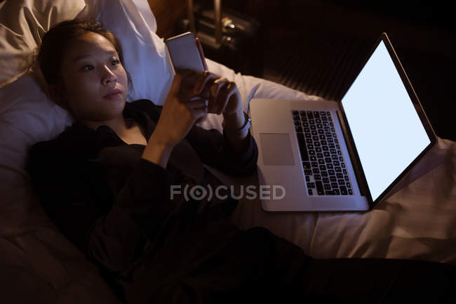Жінка використовує мобільний телефон, розслабляючись на ліжку в готелі — стокове фото