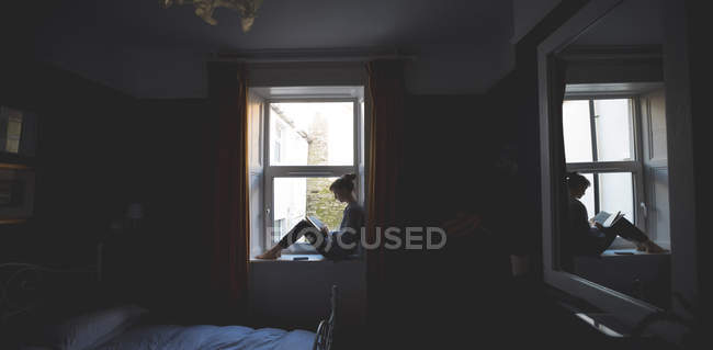 Woman reading book on window sill in bedroom at home — Stock Photo