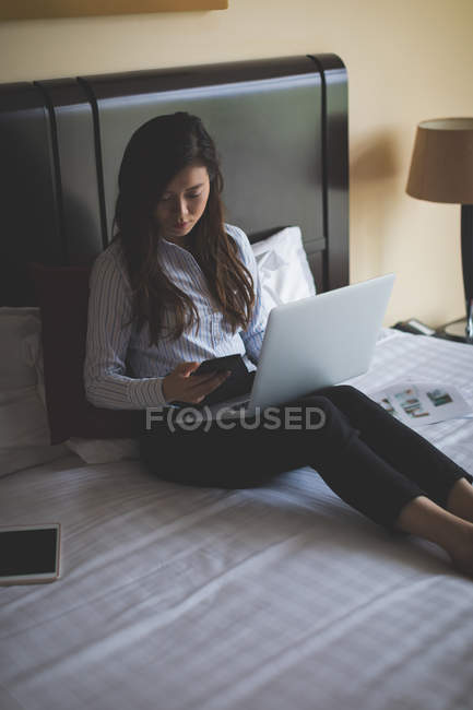 Бізнес-леді використовує свій мобільний телефон під час роботи на ноутбуці в готельному номері — стокове фото
