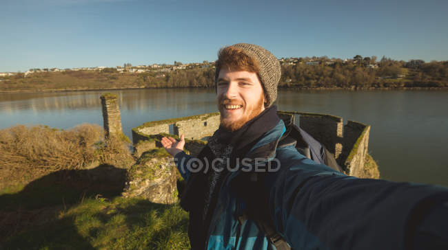 Felice escursionista maschio in piedi e in posa per selfie in rovina vicino al fiume in campagna — Foto stock