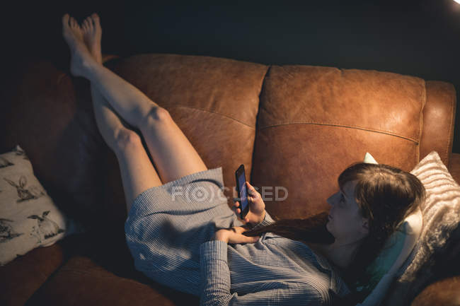 Mujer usando el teléfono móvil mientras está acostado en el sofá en la sala de estar - foto de stock