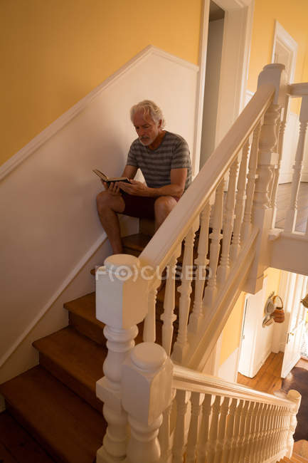 Homem idoso ativo lendo um livro nas escadas em casa — Fotografia de Stock