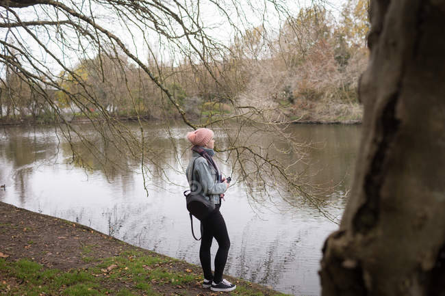 Young woman with back pack standing near river coast at park — Stock Photo