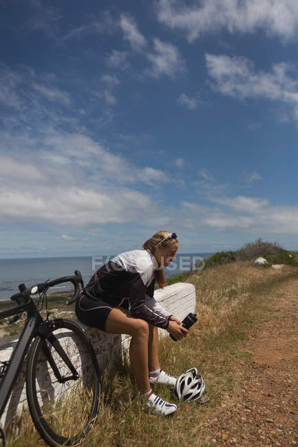 Feminino motociclista com garrafa de água sentado no banco de rocha em um dia ensolarado — Fotografia de Stock