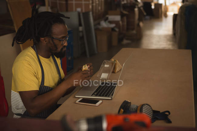 Тесляр їсть сендвіч під час роботи на ноутбуці в майстерні — стокове фото