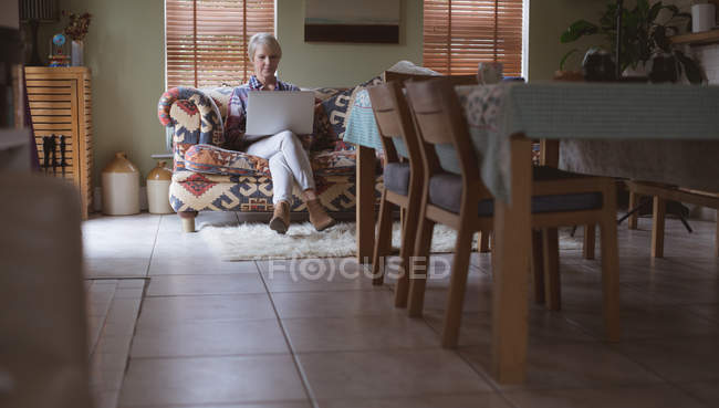 Femme mûre utilisant un ordinateur portable sur le canapé dans le salon à la maison — Photo de stock