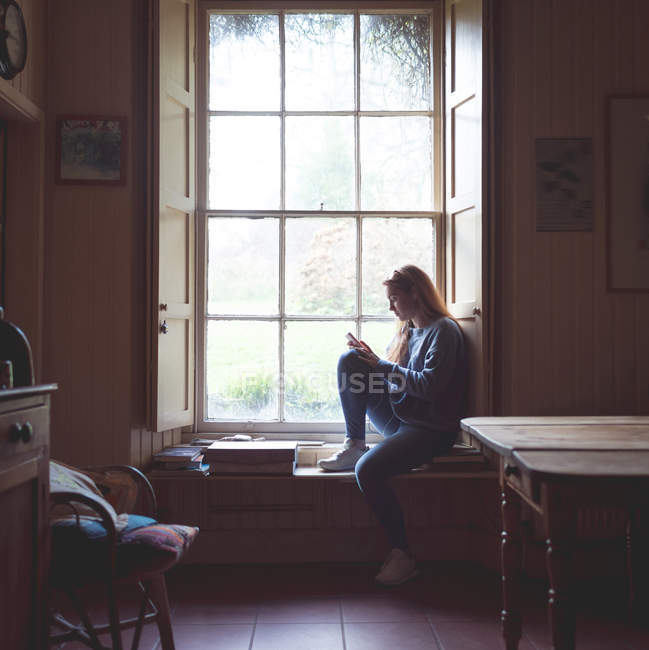 Mujer usando teléfono móvil cerca de la ventana en casa - foto de stock