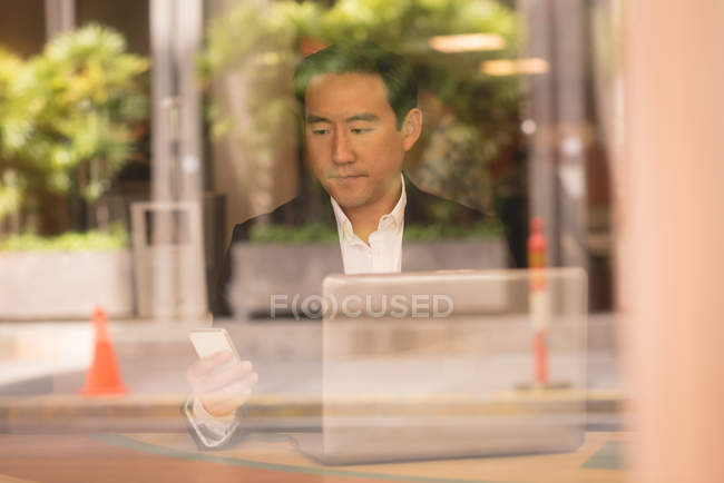 Hombre de negocios asiático utilizando el teléfono móvil en la cafetería detrás de la ventana de vidrio - foto de stock