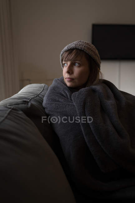 Задумчивая женщина, завернутая в одеяло на диване дома . — стоковое фото