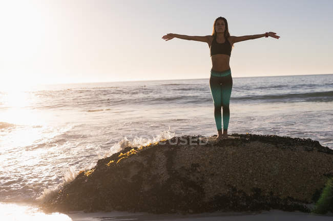 Mujer en forma realizando yoga sobre roca en la playa al atardecer . - foto de stock