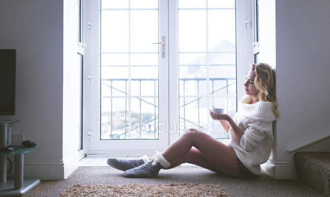 Задумчивая женщина пьет кофе у окна дома — стоковое фото