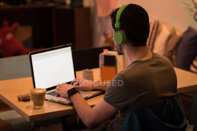 Vista trasera del hombre usando el ordenador portátil con auriculares en la cafetería . - foto de stock
