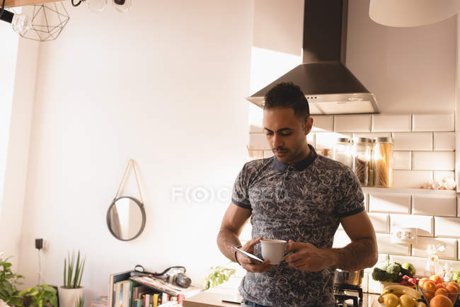 Man holding cup of coffee and smartphone in kitchen at home. — Stock Photo