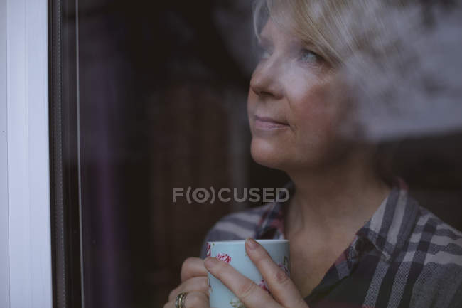 Pensativo mujer madura sosteniendo taza de café mientras mira a través de la ventana en casa - foto de stock